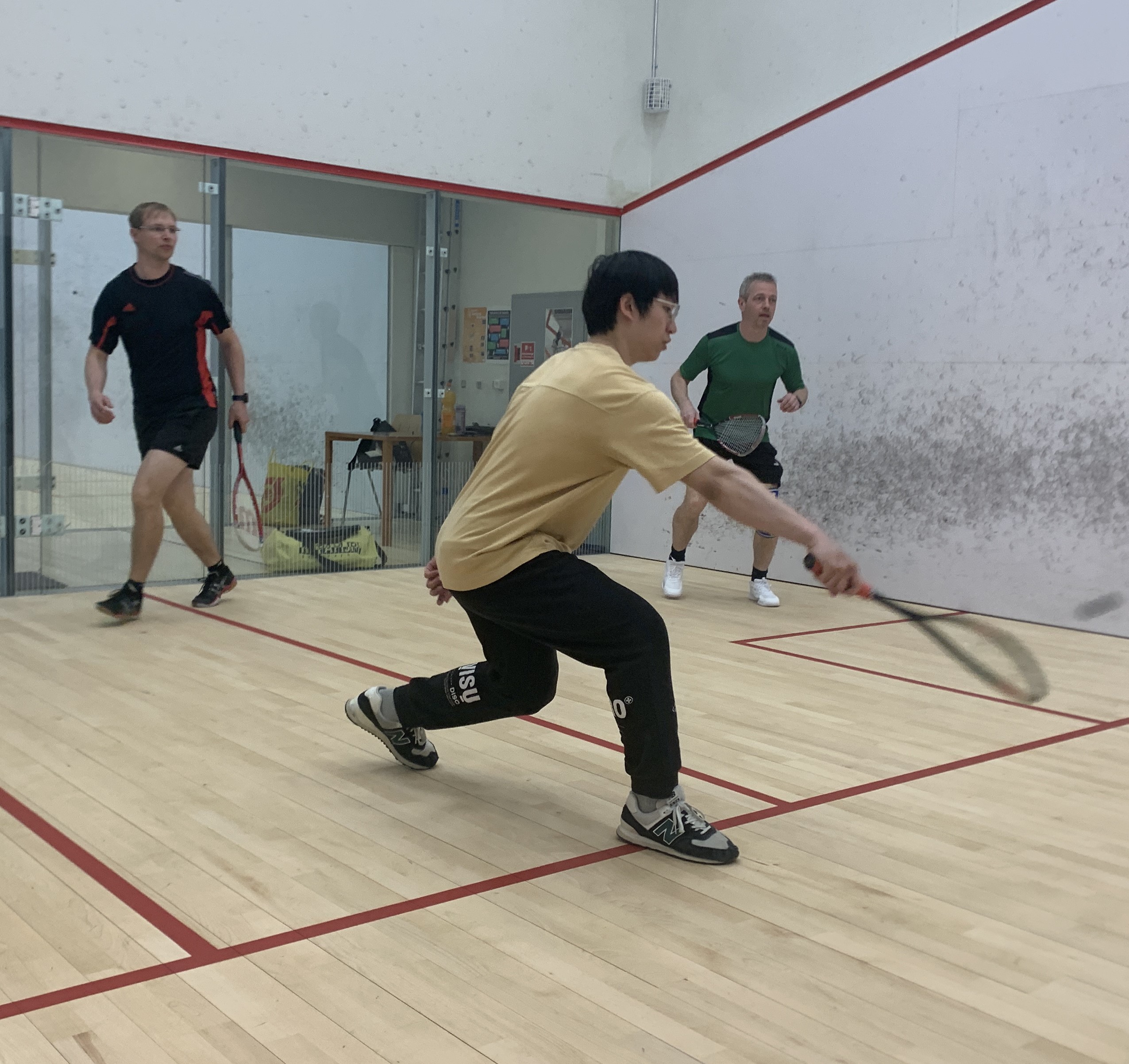 Tre spillere på en squashbane, hvor en spiller i forgrunden når frem til bolden med sin ketsjer.