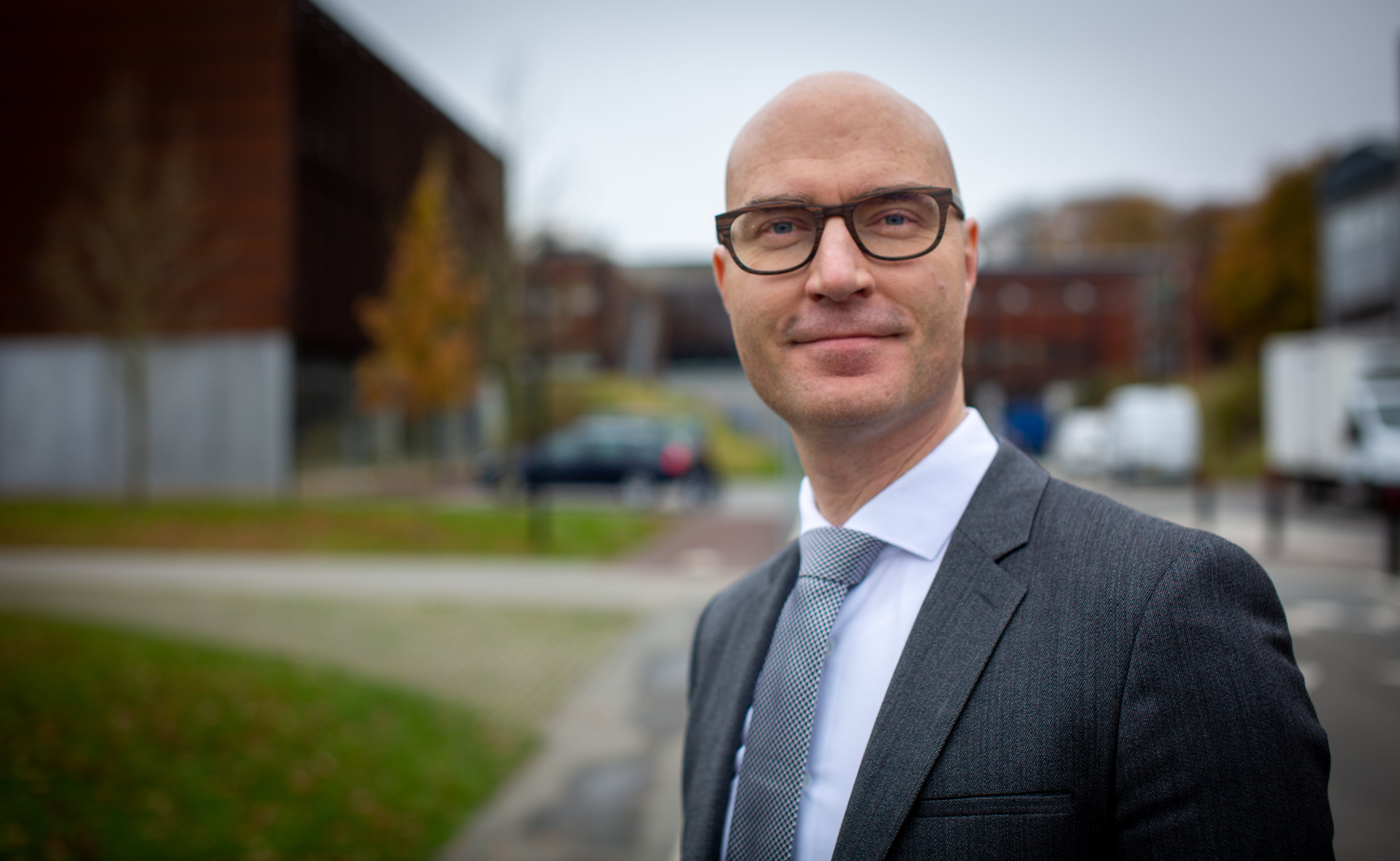 Skaldet mand med briller i jakkesæt står udendørs foran en sløret bymæssig baggrund.