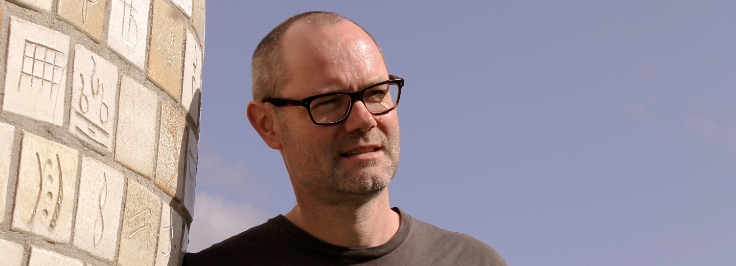 Man wearing glasses standing beside a carved stone wall outdoors under a cloudy sky.