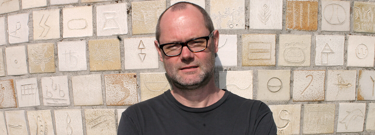 Man with glasses standing in front of a wall of engraved tiles with symbols.