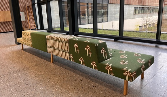 Lang bænk med unikt, naturinspireret stofdesign i grønne nuancer og mønstre af svampe og planter, placeret i en moderne bygning med store vinduer og udsigt til en grøn gårdhave.