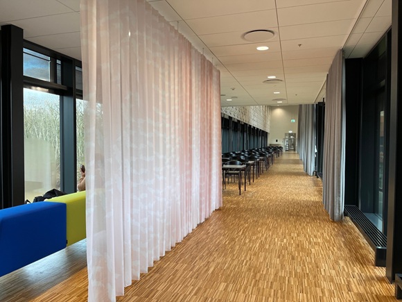 Lang gang i en moderne bygning med trægulv, sorte stole ved borde og store vinduer. Lyserøde og grå gardiner deler rummet, og en farverig bænk står ved vinduet.