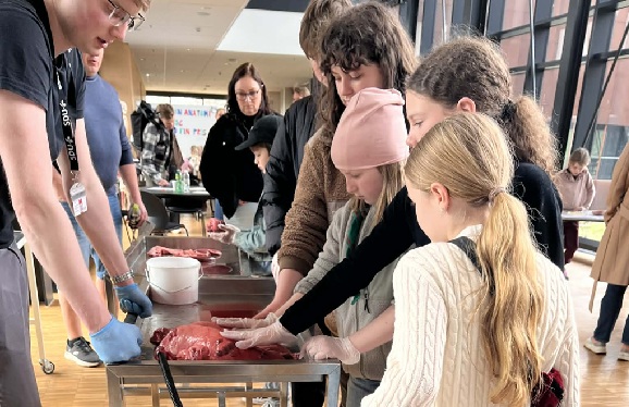 Børn deltager i en hands-on videnskabsaktivitet, hvor de undersøger organer på et bord sammen med en formidler iført handsker, i et lyst læringsmiljø med andre deltagere i baggrunden.