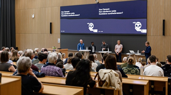 Paneldebat med forskere og en vært på en scene foran et fyldt auditorium under en videnskabelig konference, med storskærm og præsentation i baggrunden.