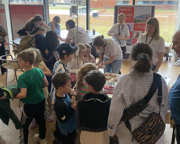 Børn og familier deltager i en hands-on aktivitet ved en formidlingsstand, hvor frivillige guider dem gennem eksperimenter og læring.