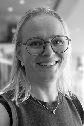 Sort-hvidt portræt af en smilende kvinde med briller og lyst skulderlangt hår indendørs.