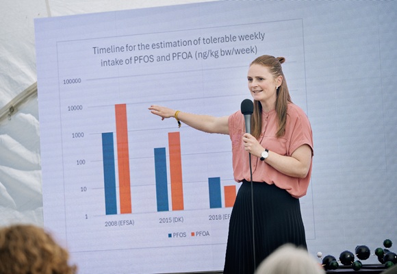 A woman gives a presentation with a microphone in front of a large diagram showing the development of tolerable weekly intake of PFOS and PFOA. She points to the bar chart and addresses an audience during an outdoor event.