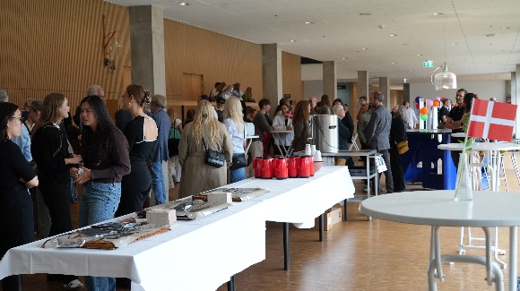Studerende og deltagere netværker ved en reception eller faglig begivenhed i en moderne bygning med træpaneler og store vinduer. Forreste borde er dækket med kaffekander, kage og informationsmateriale. Et lille bord med danske flag ses i højre hjørne.