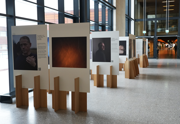 Photo exhibition in a modern building with large glass windows and snow outside. The exhibition is titled "Do You See What I See" and features photographs mounted on white panels with wooden stands. The images and accompanying texts are arranged in a long row in a bright and open hallway.