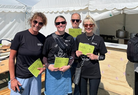 Four smiling people wearing black SDU T-shirts hold green information cards in front of a white tent on a sunny day. They are taking part in an outdoor event, possibly related to SDU (University of Southern Denmark).