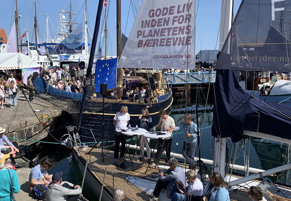 A panel of speakers stands on a boat in a busy harbour, surrounded by spectators on a sunny day. A large sail carries the message 'Gode liv inden for planetens bæreevne' (Good lives within the planet’s limits). The event appears to focus on sustainability and wellbeing.