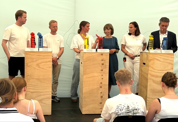 Et panel med seks personer står bag podier med farvede vandflasker under en debat foran et siddende publikum. Arrangementet foregår indendørs under lyse forhold, og emnet synes at være relateret til miljø eller sundhed.