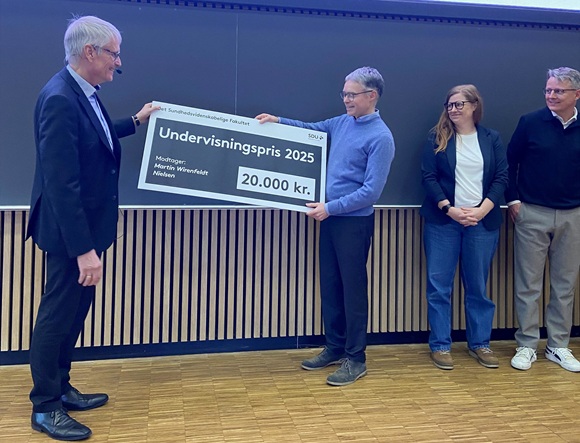 Fire personer står foran en tavle i et auditorium, hvoraf én modtager Undervisningspris 2025 fra Det Sundhedsvidenskabelige Fakultet ved SDU. Personen i midten holder et stort symbolsk checkskilt på 20.000 kr. med sit navn, Morten Wernefelt Nielsen. De øvrige personer ser glade ud og fejrer øjeblikket.