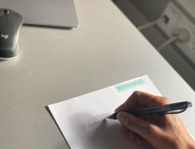 En person holder på en kuglepen og er i gang med at skrive noget på et hvidt stykke papir. 