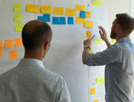 To mænd står foran et whiteboard og kigger på post-its, som hænger på whiteboardet.