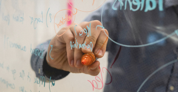 En hånd der holder en orangefarvet tusch – og skriver på en glastavle.