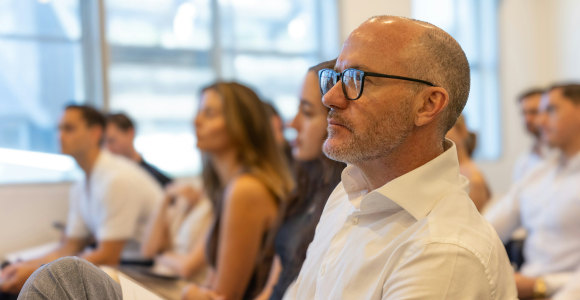 Deltagere til et arrangement sidder, lytter og kigger ligefrem.